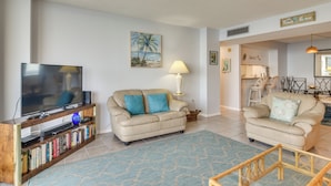 A living room with a flat-screen TV, a beige sofa, a coffee table, and a bookshelf. - Dolphin & Manatee Viewing From 15th Floor & Pool! (Clearwater Beach)