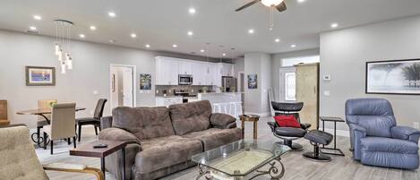 A spacious living room with a brown leather sofa, a glass coffee table, and a blue armchair. There is a kitchen in the background with white cabinets and a ceiling fan.