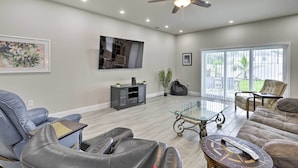 A spacious living room with leather sofas, a glass coffee table, a ceiling fan, and a large TV mounted on the wall. - Palm Bay Home w/ Patio & Yard - 12 Mi to Beach! (Palm Bay)