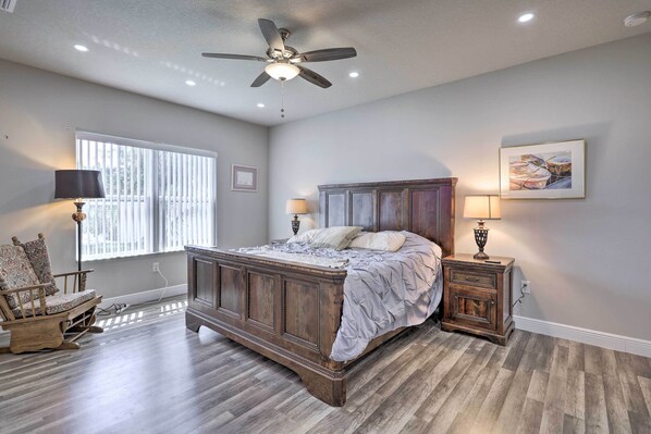 A bedroom with a wooden bed, a ceiling fan, a window with blinds, a nightstand with a lamp, and a chair.
