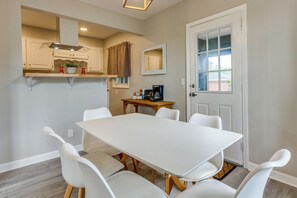 A dining area with a white table and chairs, a kitchen with beige cabinets, a window with curtains, and a door with a glass panel.