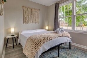 A neatly arranged bedroom with a bed, bedside lamps, a macrame wall hanging, and a large window with a view of the outside.