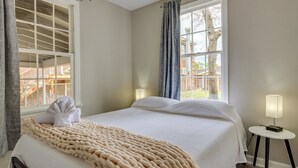 Ein ordentlich bezogenes Bett mit einer beigen Decke, einem weißen Kissen und einem Handtuch darauf. Ein Fenster bietet Blick auf eine Holzterrasse und einen Baum. Ein kleiner Tisch mit einer Lampe steht neben dem Bett.