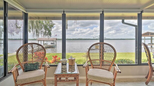 Uma área de estar coberta ao ar livre, com cadeiras de vime e uma mesa, com vista para um lago com um píer e uma área gramada.