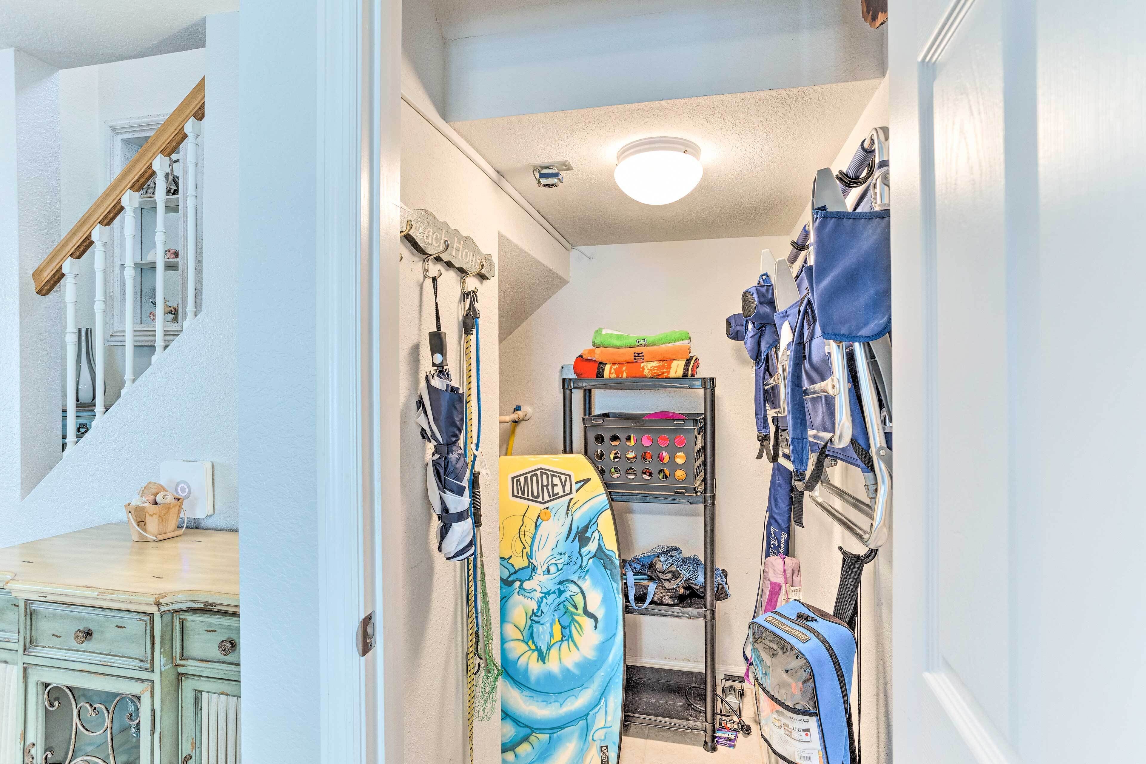 A narrow hallway with a staircase on the left, a surfboard leaning against the wall, and various bags and gear on shelves.