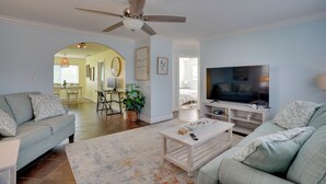 Ein Wohnzimmer mit Deckenventilator, Fernseher, zwei Sofas, einem Couchtisch und einem Essbereich im Hintergrund.