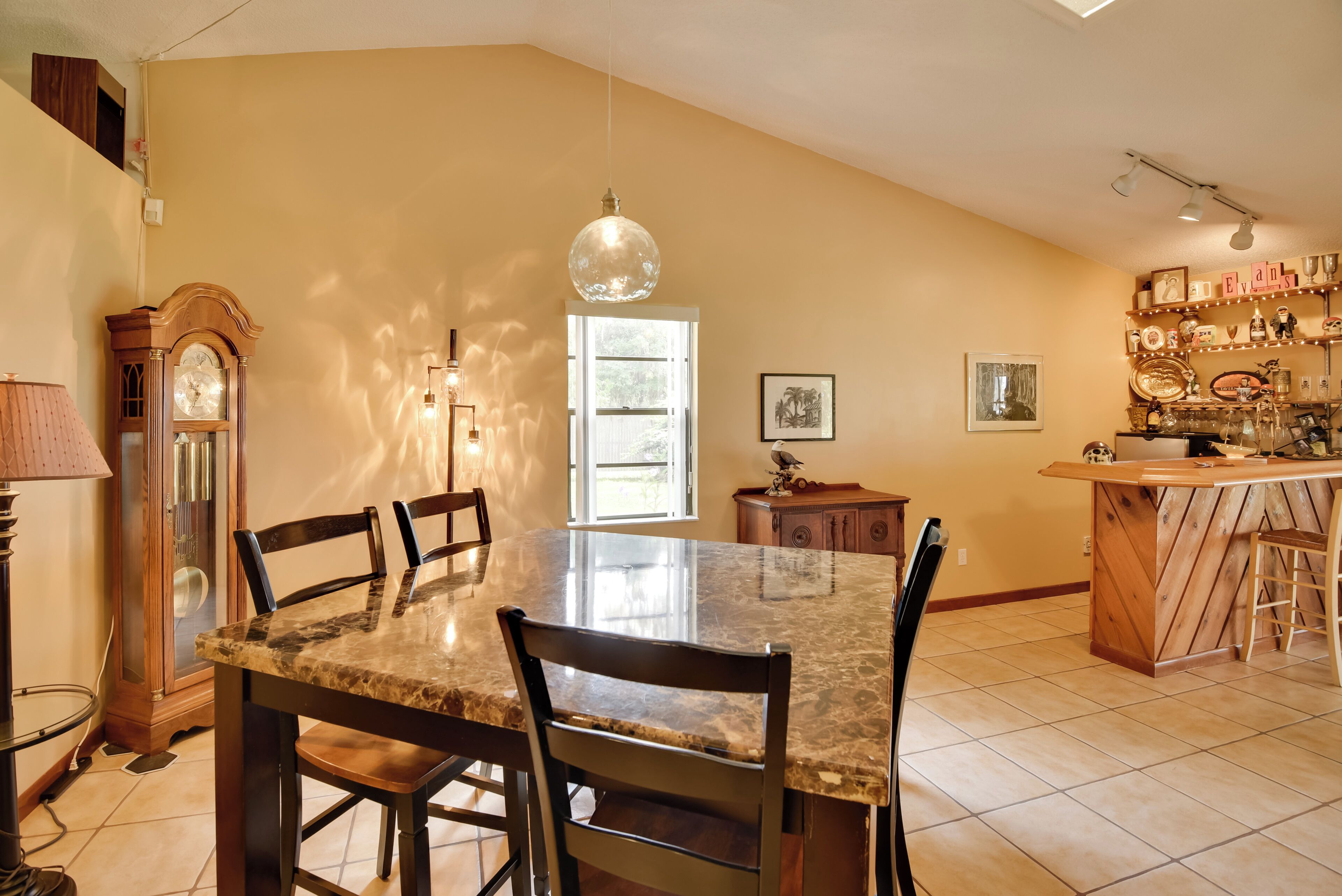 A dining area with a marble table and chairs, a wooden clock, and a kitchen area with a wooden bar and shelves.