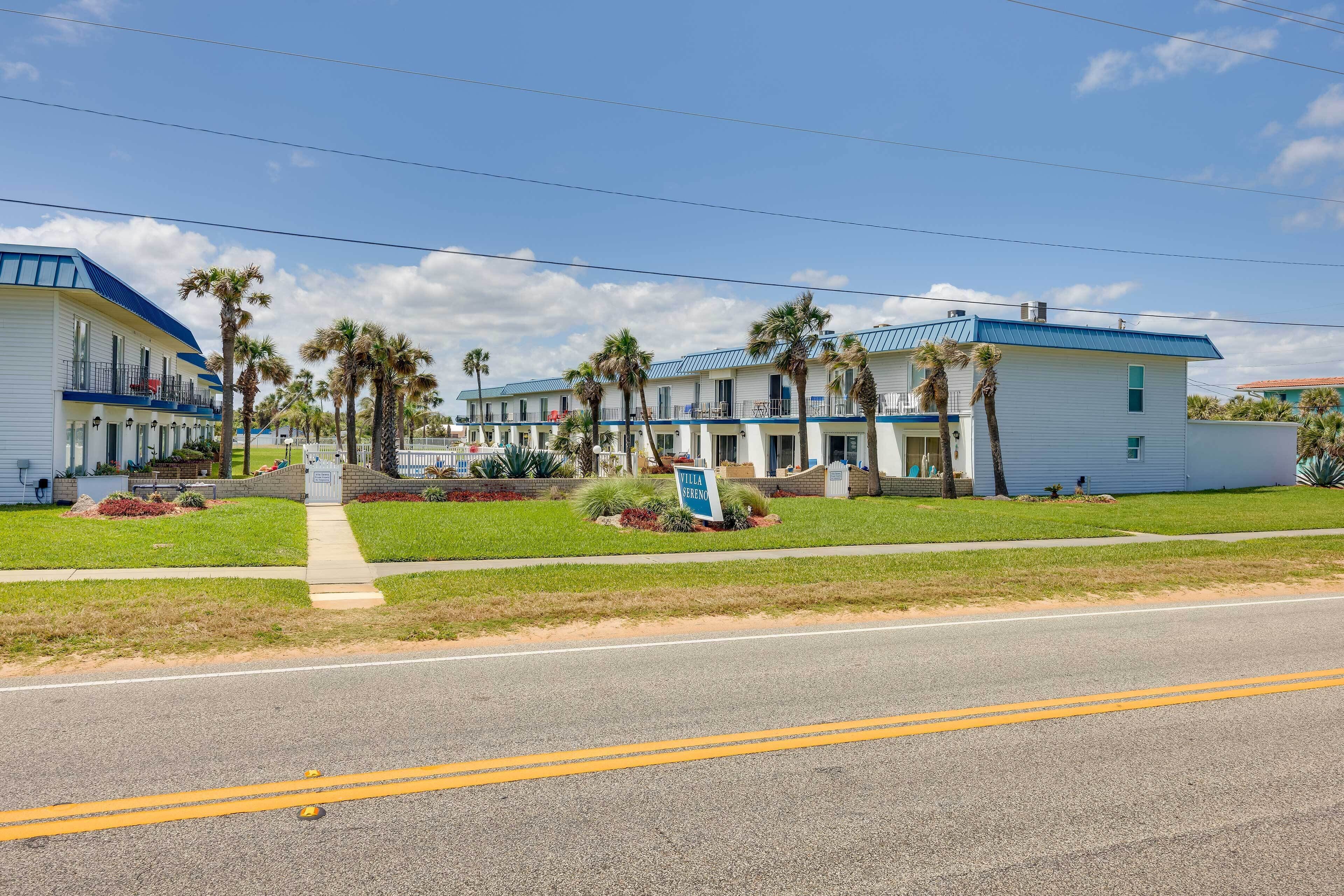 A row of motel buildings with a blue roof, surrounded by palm trees and a clear sky.
