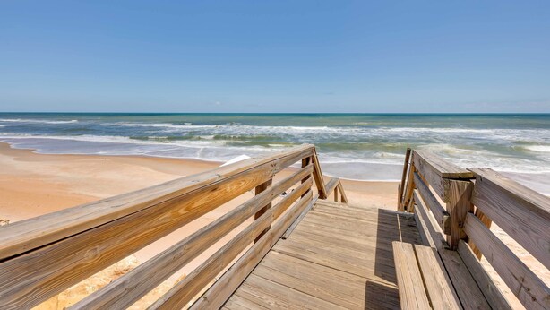 Ein hölzerner Steg führt zu einem Strand, wo Wellen auf das Ufer branden.