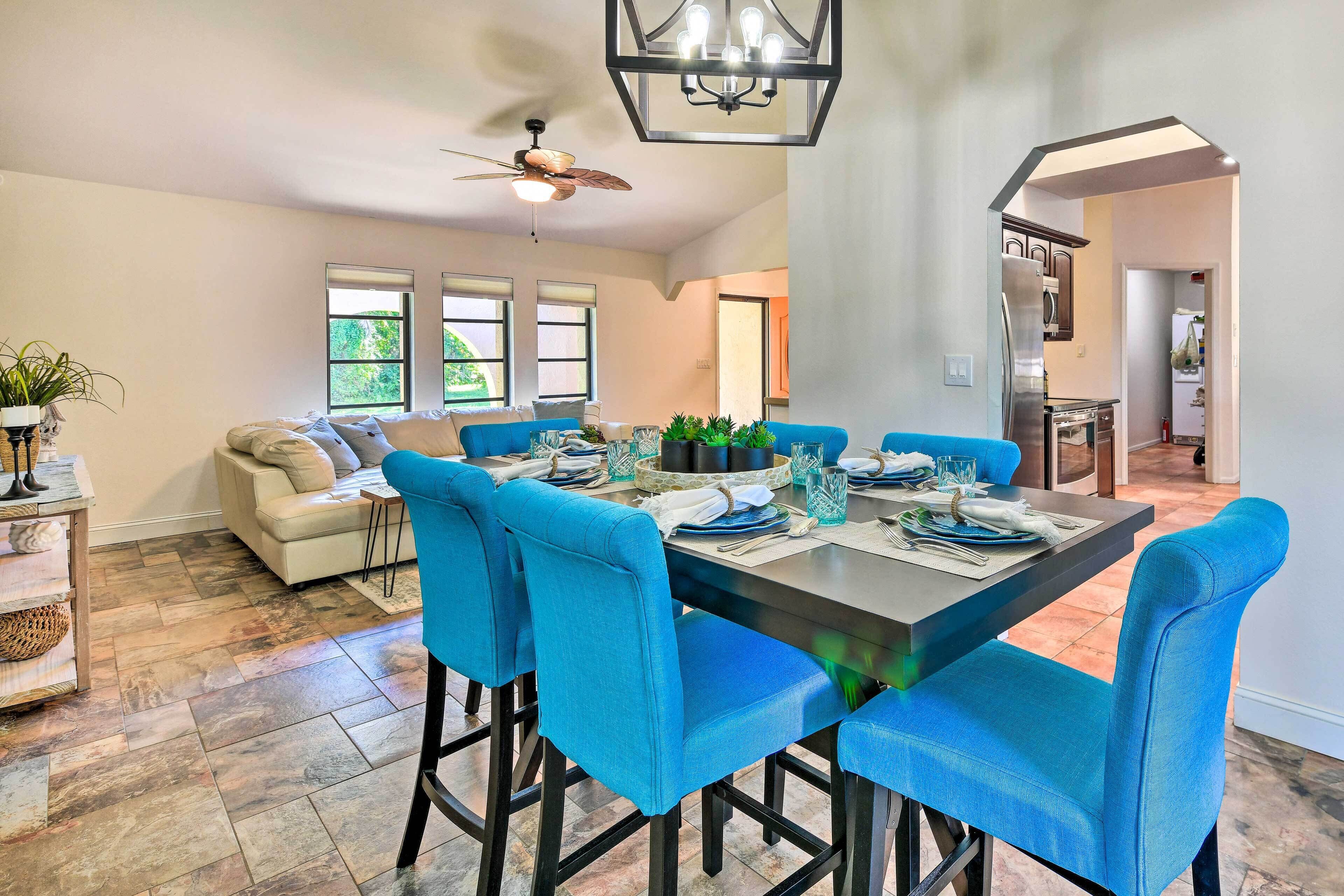 Un coin repas avec une table dressée pour six personnes, des chaises rembourrées en tissu bleu, un ventilateur de plafond et une vue sur la cuisine par une porte ouverte.