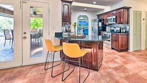 A kitchen with a central island, two yellow bar stools, wooden cabinets, and a view into the dining area. - Port Charlotte Retreat w/ Heated Pool & Spa! (Port Charlotte)