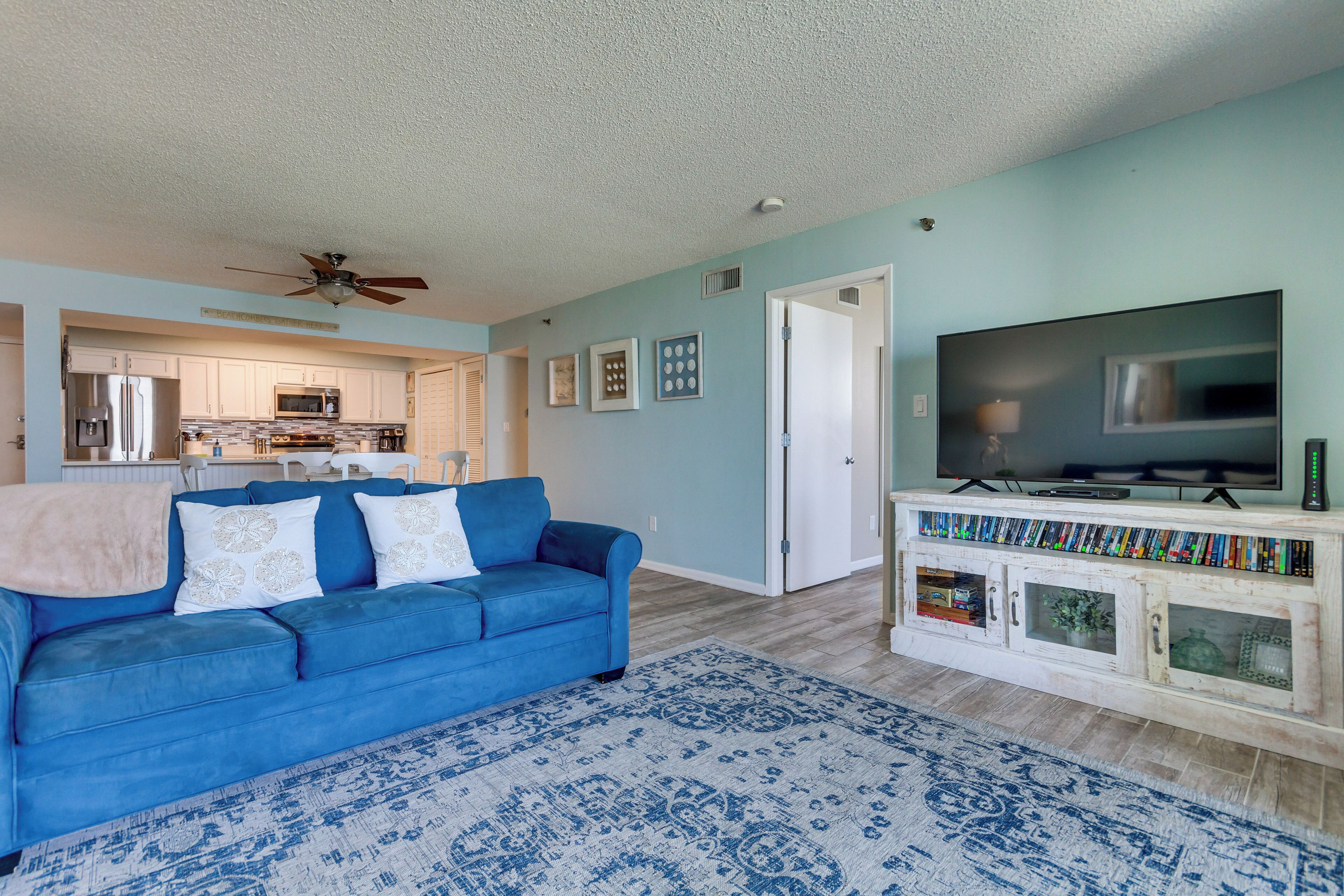A living room with a blue sofa, a patterned rug, a television on a cabinet, and a kitchen in the background.