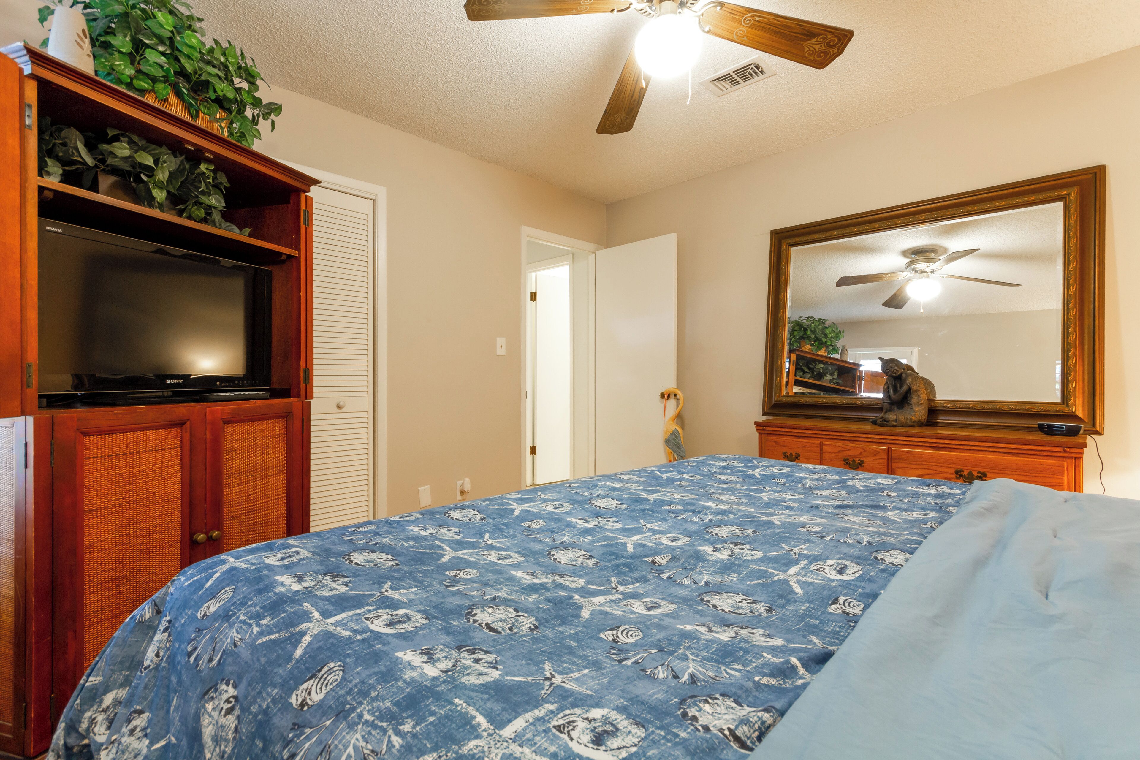 A bedroom with a bed, a television, a ceiling fan, and a wooden cabinet with plants.