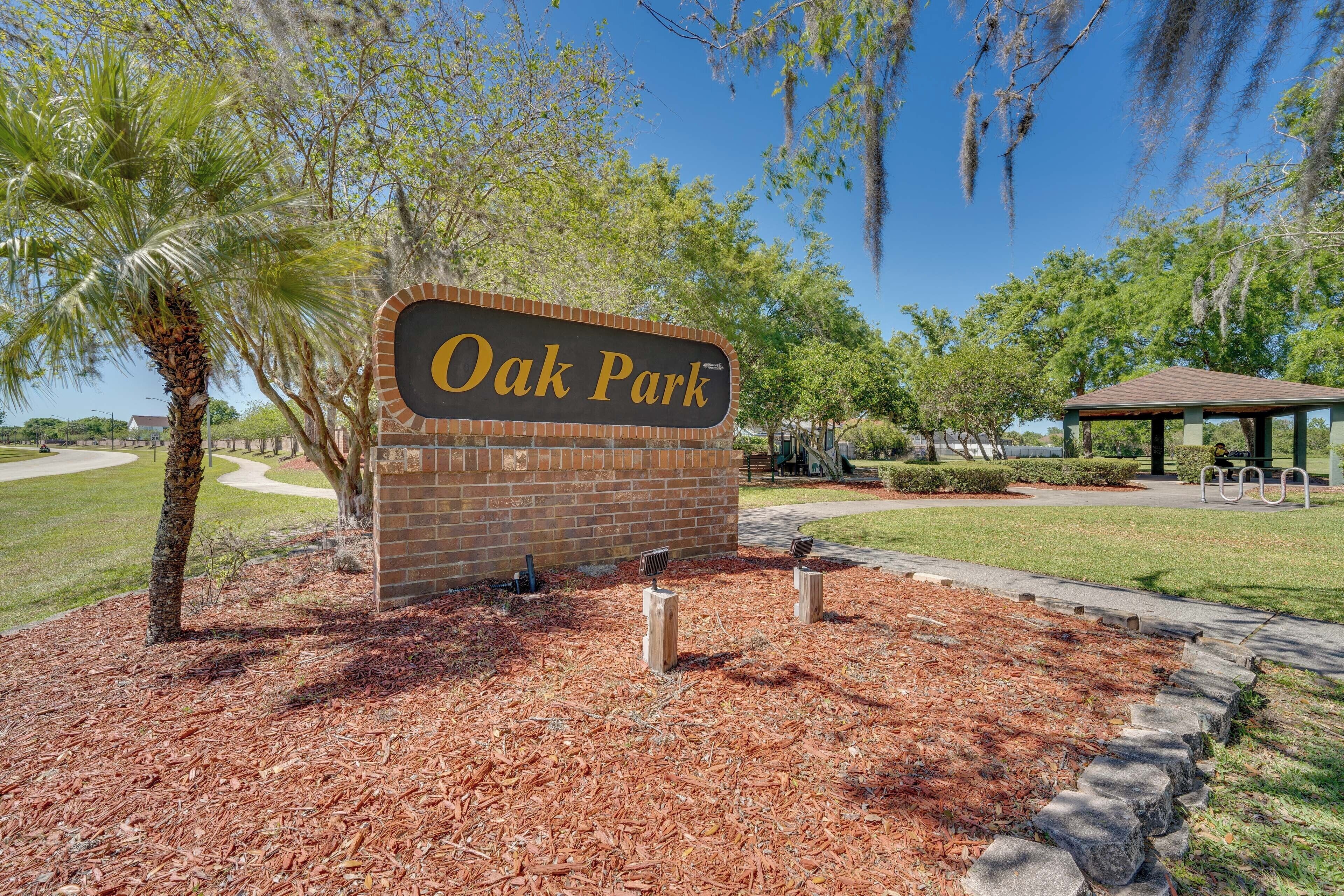 Schild für Oak Park mit Ziegelunterbau und Holzdecke, umgeben von Mulch, einer Palme und einem Gazebo im Hintergrund.