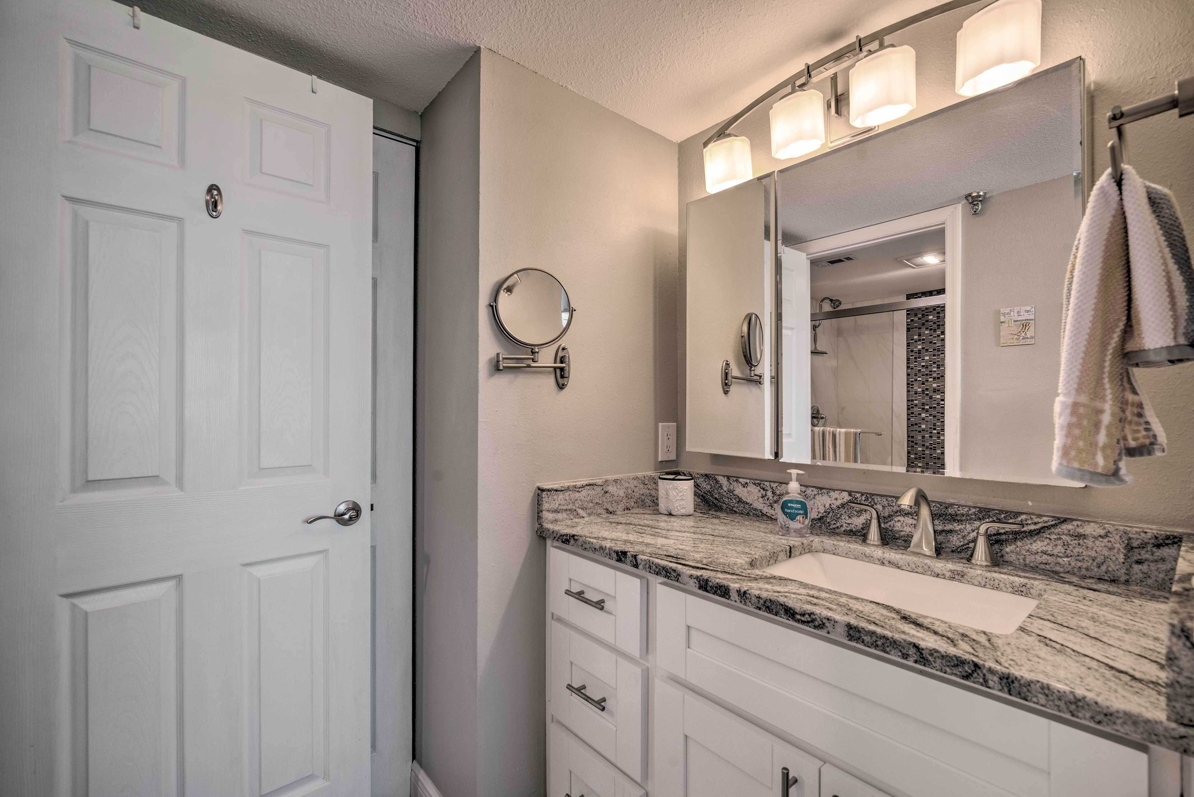A bathroom with a white door, a marble sink, a large mirror with lights, and a towel hanging on the wall.