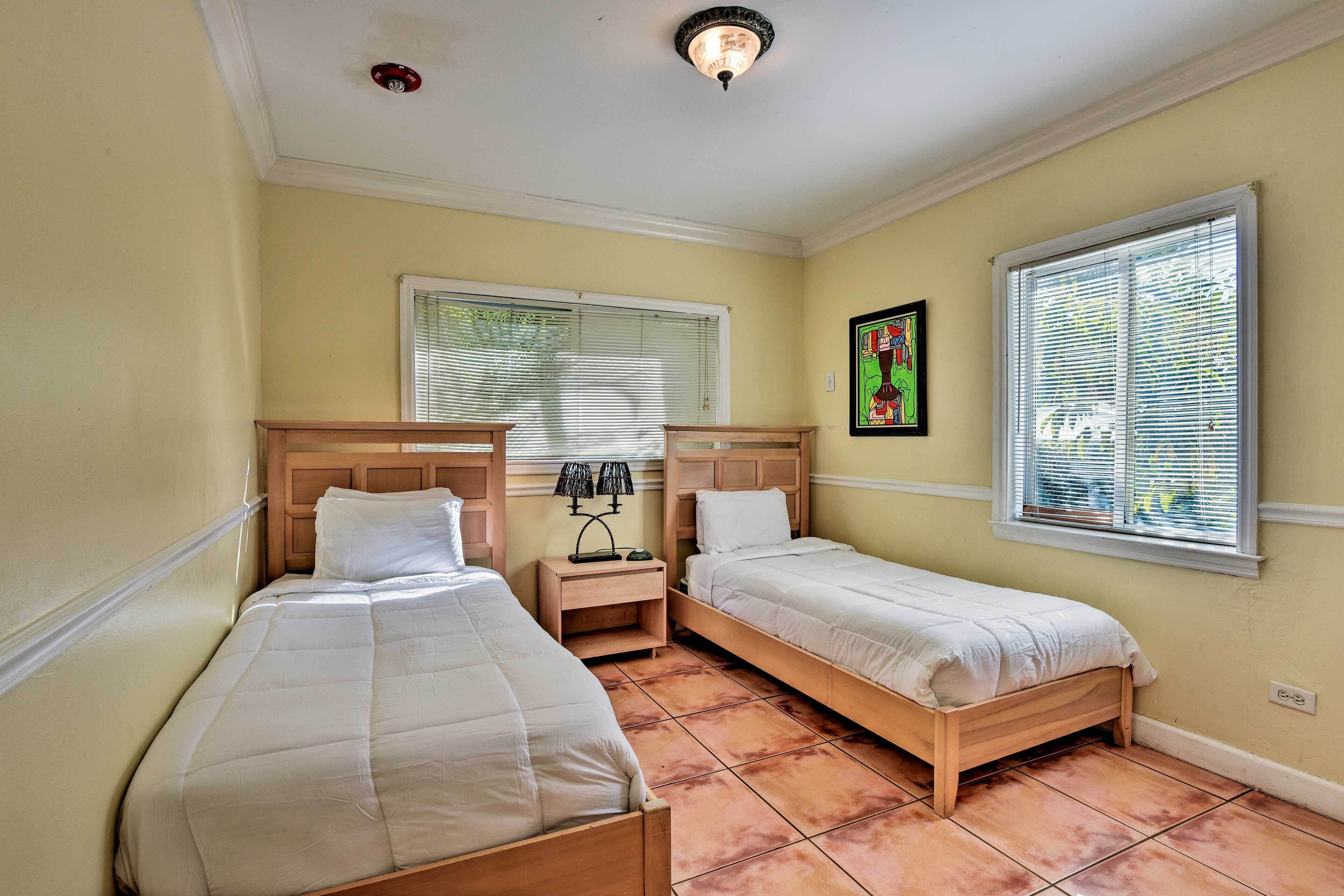 A room with two single beds, a nightstand with lamps, a window with blinds, and a ceiling light fixture.