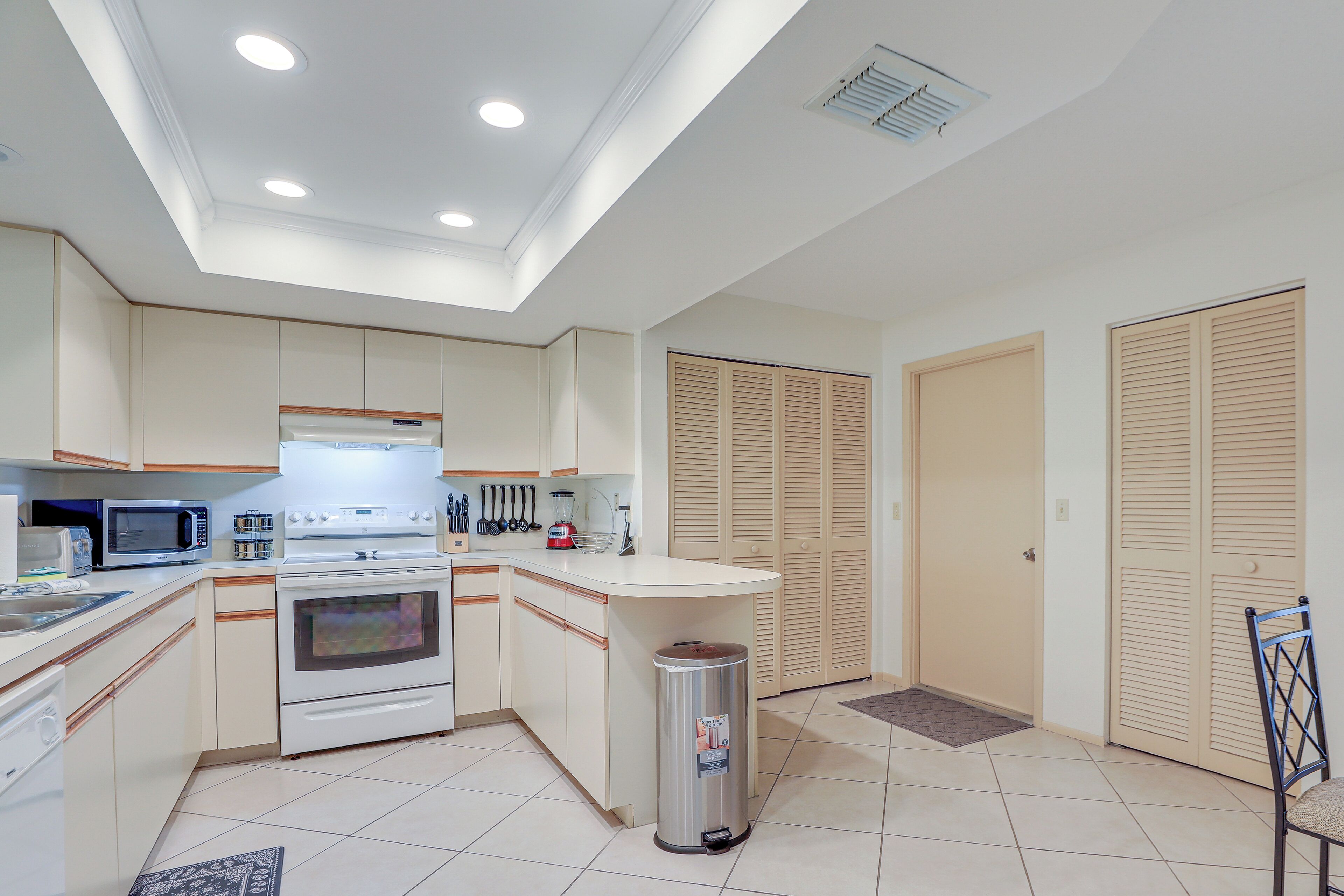 A modern kitchen with white cabinets, a built-in oven, a microwave, and a sink. There are wooden cabinets above the oven and a trash can in the corner.