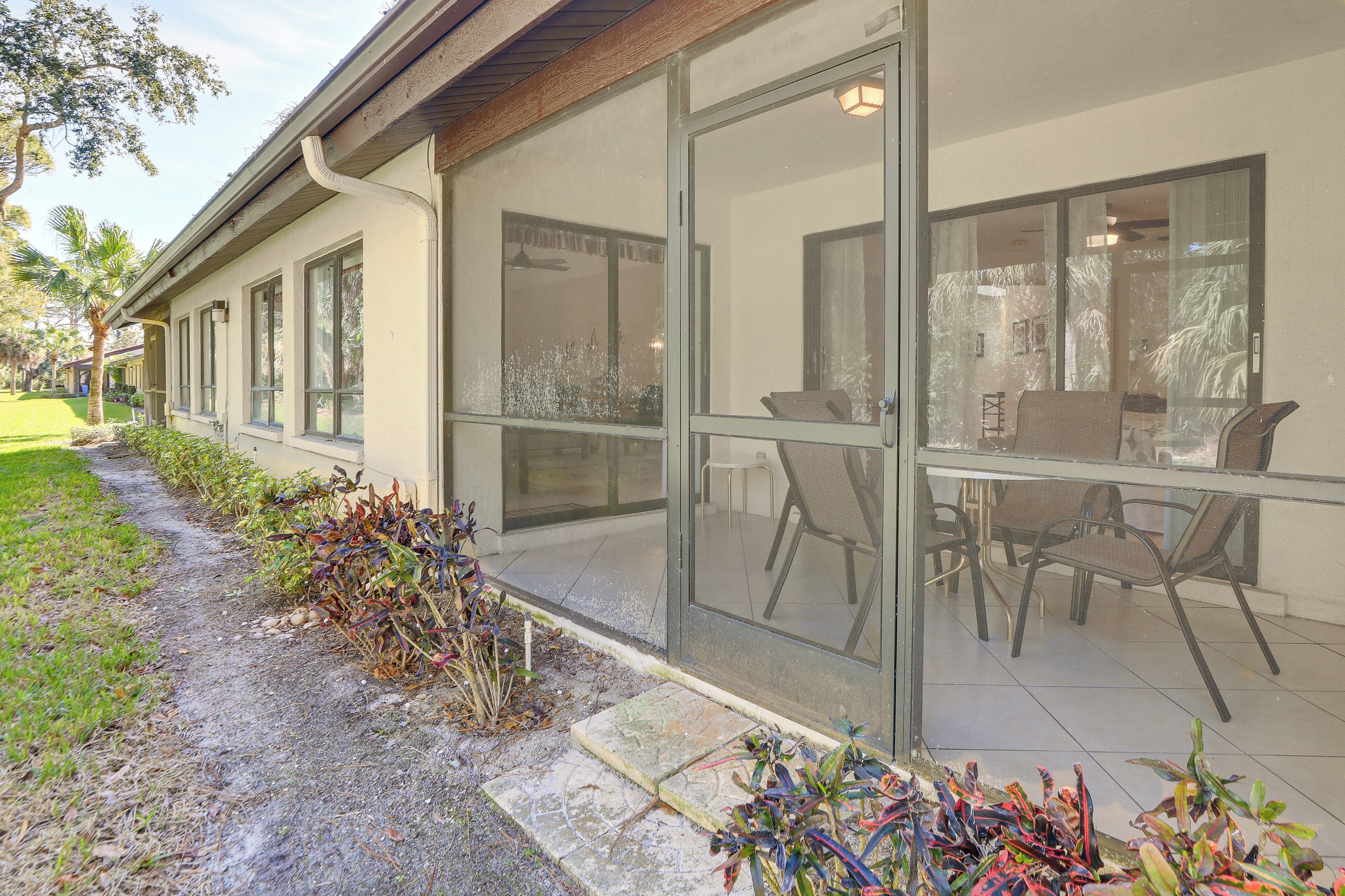 A building with large glass windows, a covered patio area with chairs, and a well-maintained garden with plants and a pathway.