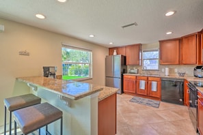 A kitchen with granite countertops, wooden cabinets, a refrigerator, dishwasher, and a window providing a view of the outdoors. - Riverfront Home w/ Putting Green & Boat Dock! (New Port Richey)