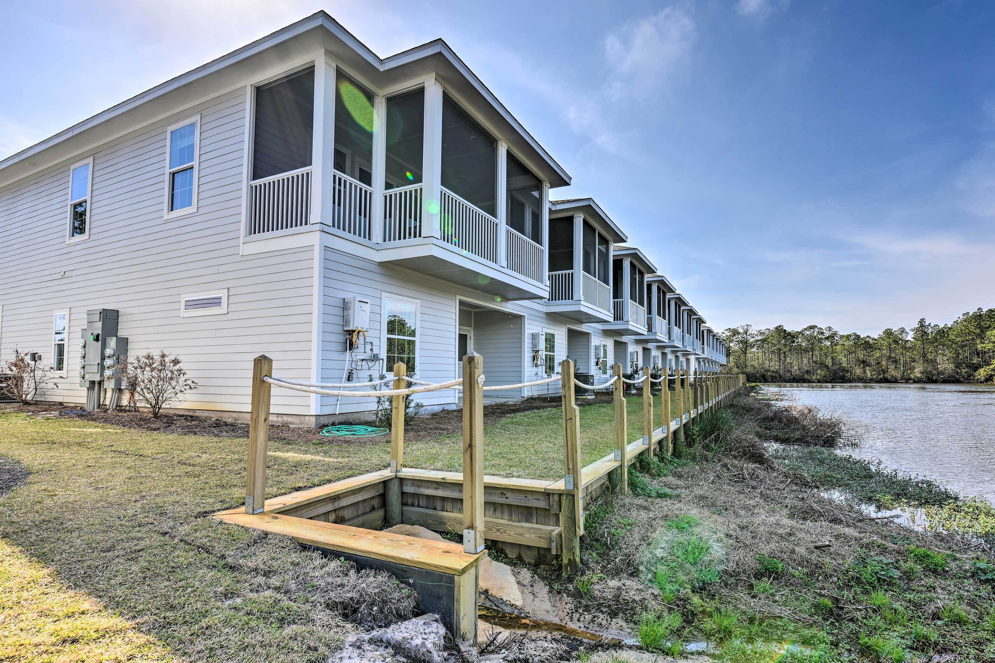 A row of waterfront houses with balconies, a wooden dock, and a grassy area.
