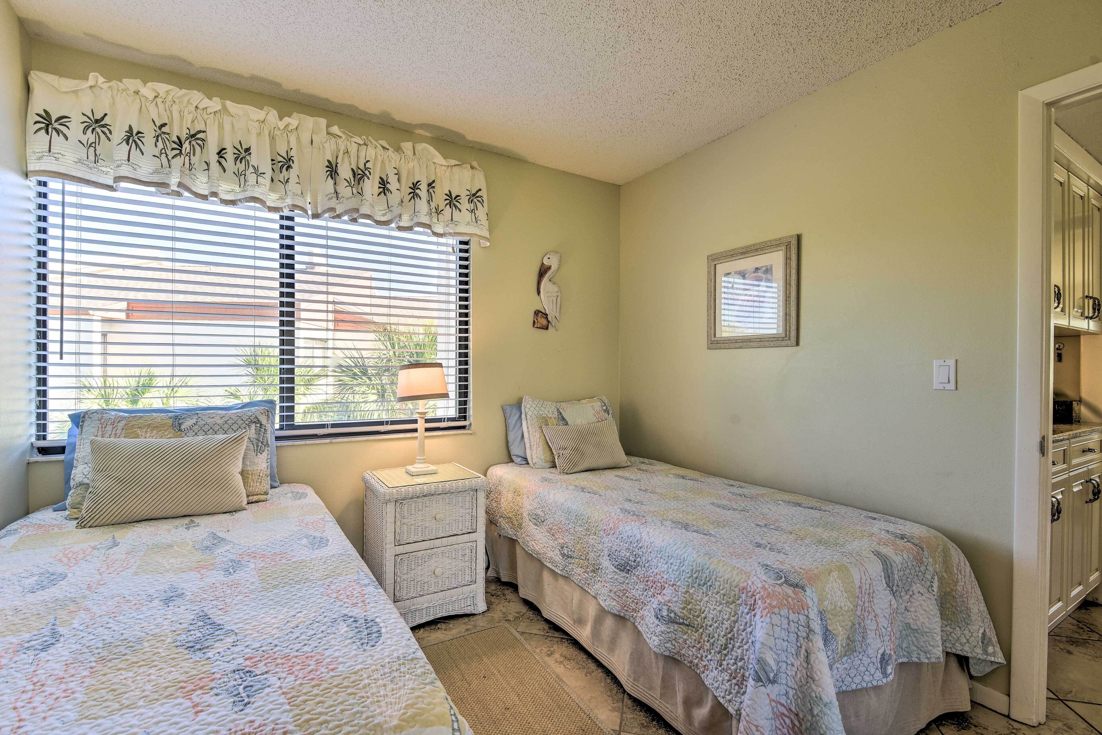 Une chambre avec deux lits simples, une table de chevet avec une lampe, une fenêtre avec des stores, un élément décoratif fixé au mur et une porte sur le côté droit.