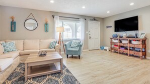 A living room with a beige sofa, a blue armchair, a coffee table, a wooden cabinet, and a TV. - Private & Picturesque Escape on Lake Henry! (Lake Placid)