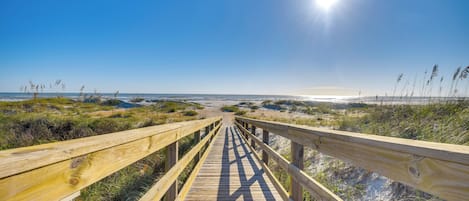 Un paseo marítimo de madera que lleva a una playa con cielo despejado y sol brillante.