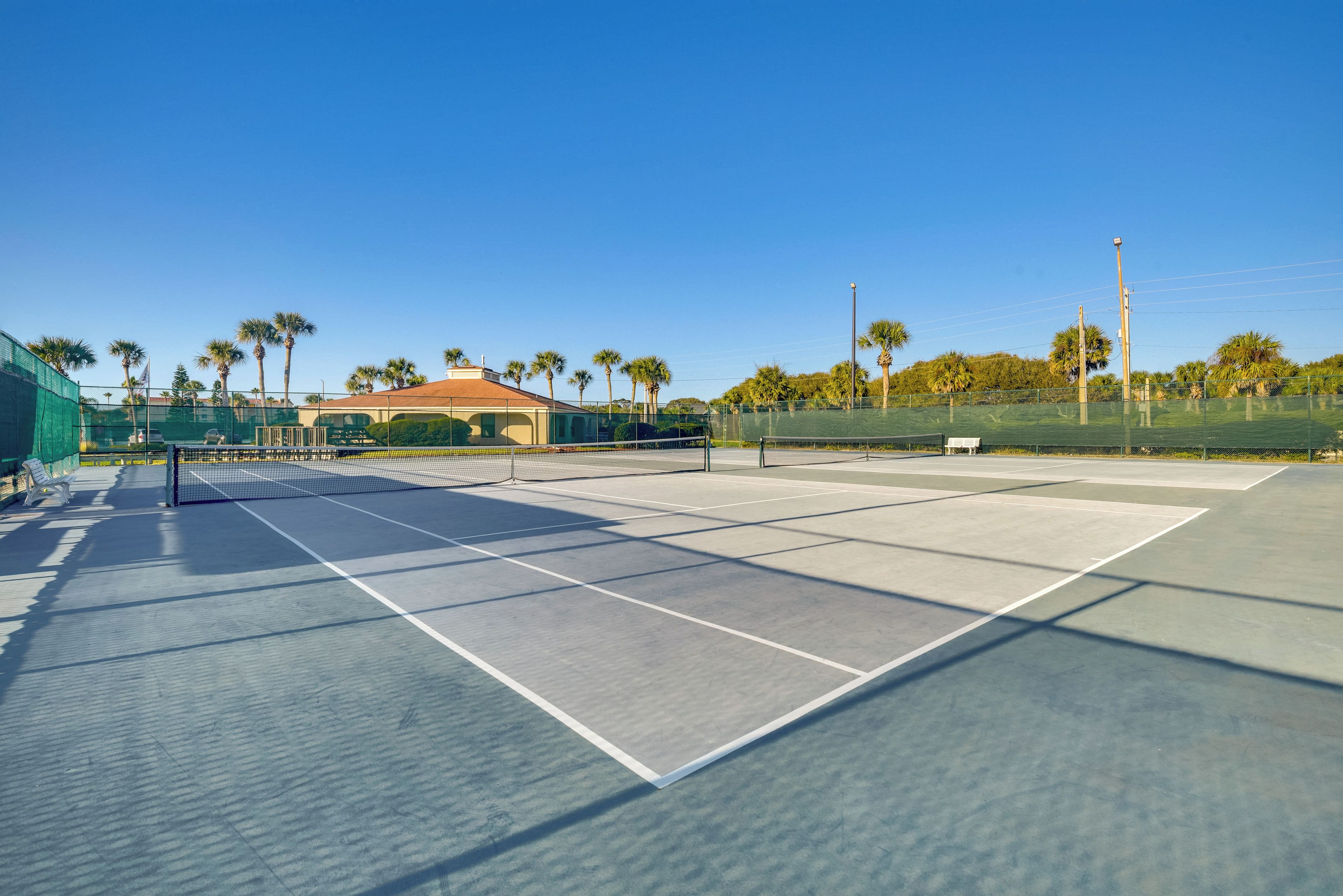 Un court de tennis en plein air, doté d’un filet, entouré de palmiers et avec un bâtiment en arrière-plan.