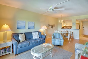 A living room with a blue sofa, a glass coffee table, and a dining area with striped chairs. - St. Augustine Condo Near Flagler College & Beach! (St. Augustine)