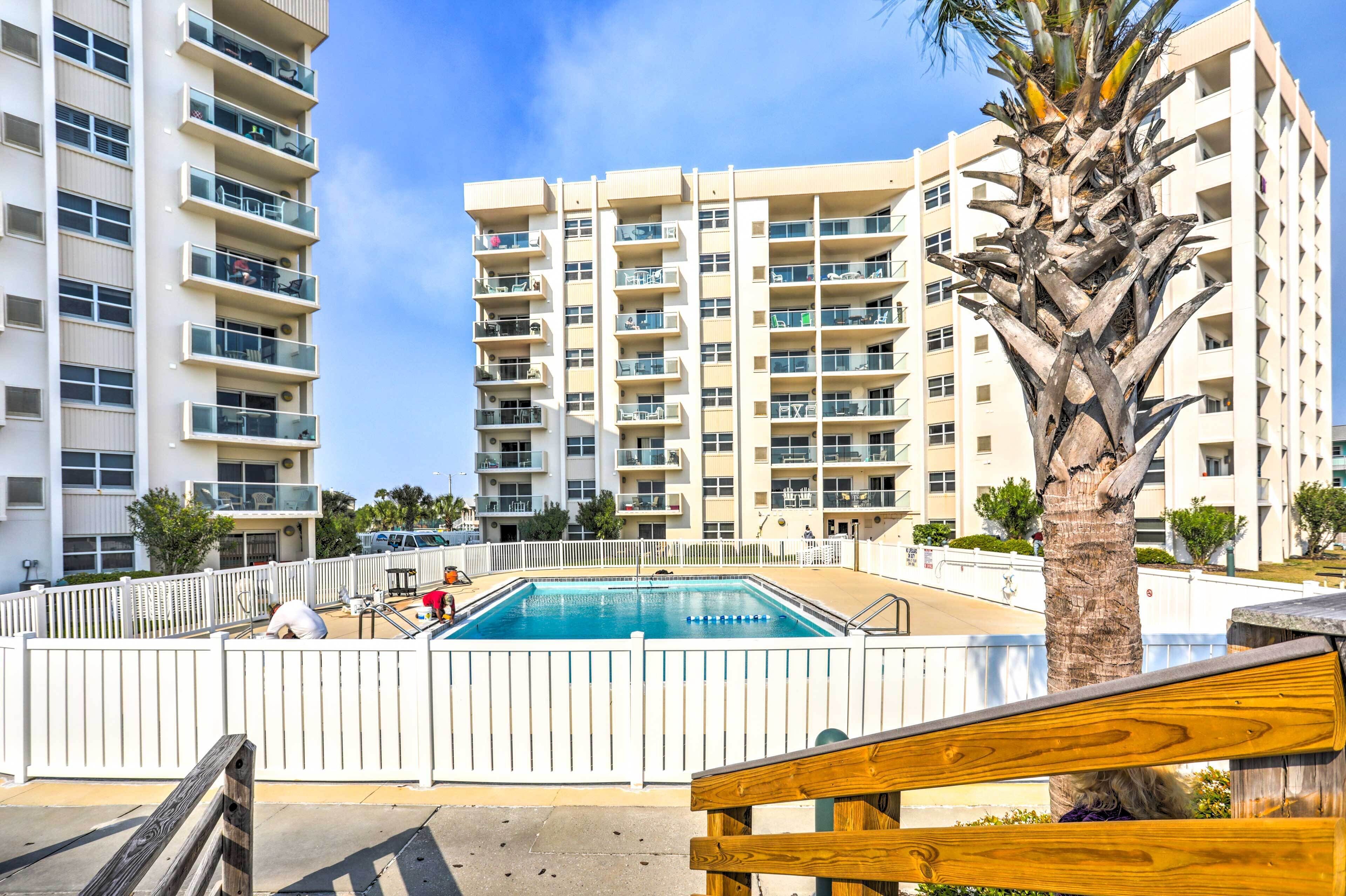 Zona de piscina con cerca blanca, una palmera y un edificio de varios pisos al fondo.