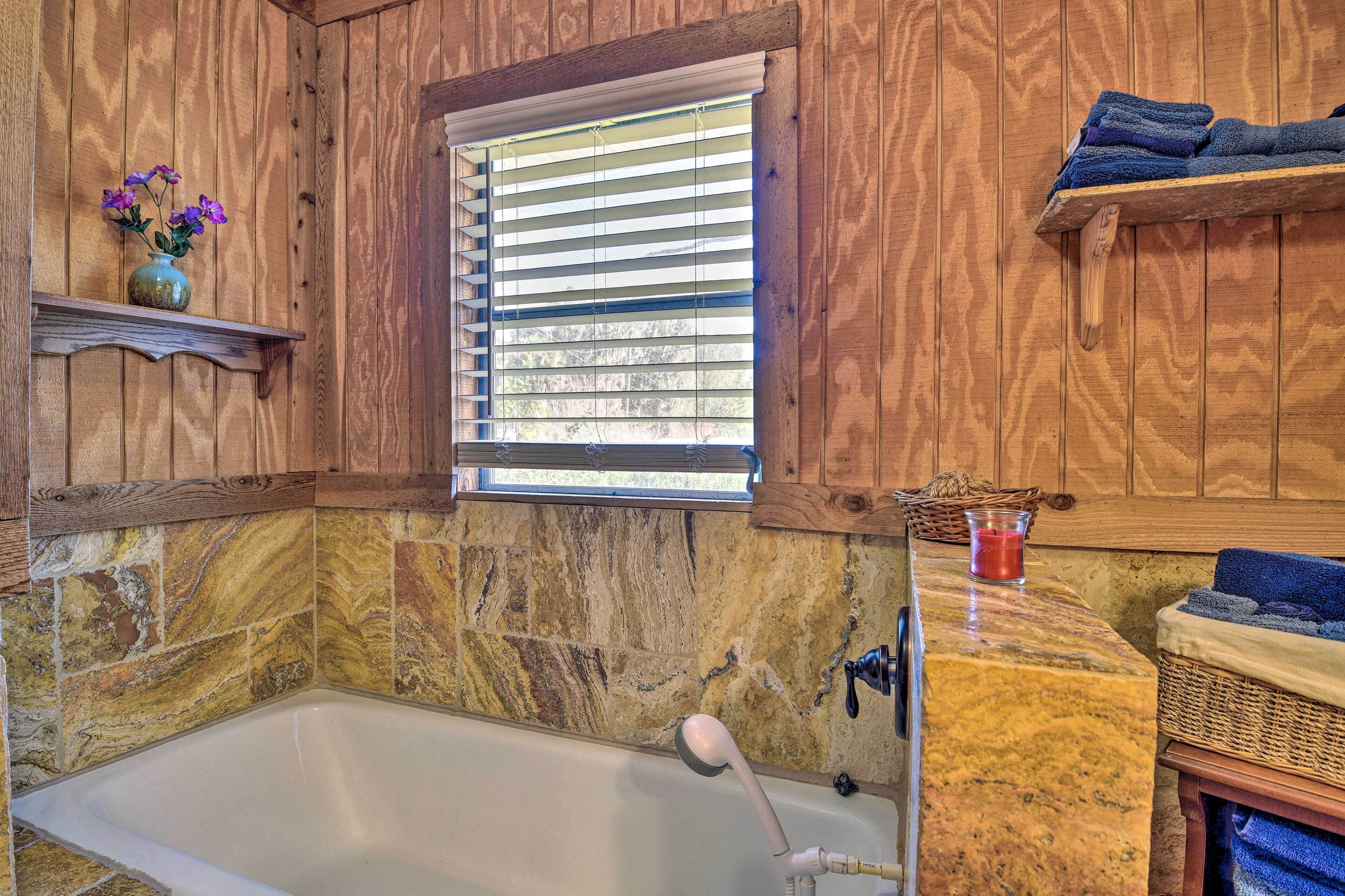 A bathroom with a stone wall, a bathtub, a window with blinds, and a shelf with towels.