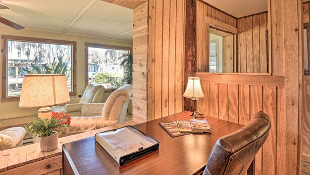 A wooden-paneled room with a desk, two lamps, a chair, and a window with a view of trees. - St Johns River Canal Home w/ Grill! (Crescent City)