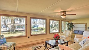 A living room with large windows, a ceiling fan, and a view of a backyard with a tree and a house. - St Johns River Canal Home w/ Grill! (Crescent City)