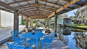 A covered outdoor patio with a wooden deck, blue metal chairs, and a table with a lantern. - St Johns River Canal Home w/ Grill! (Crescent City)
