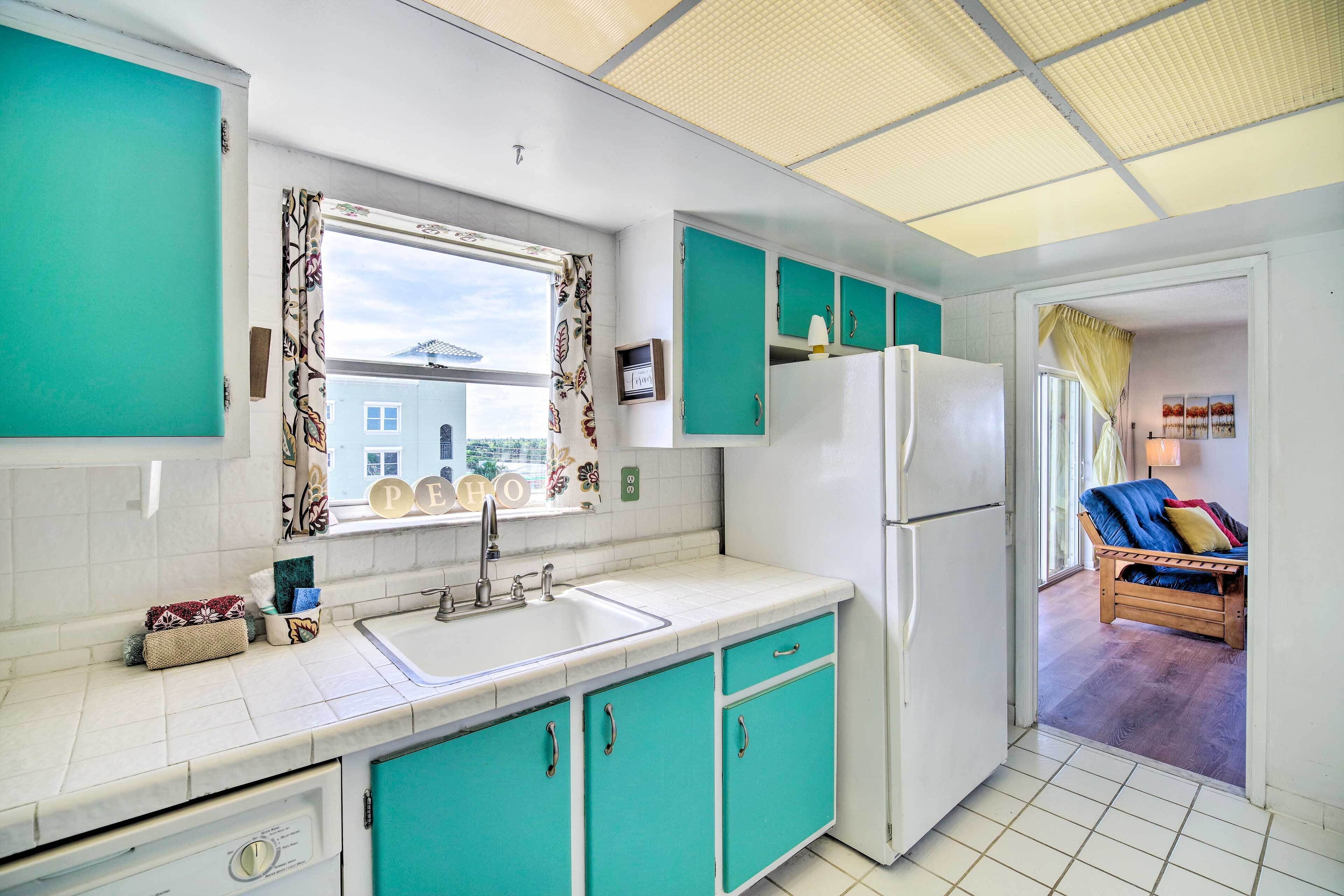 Una cocina con armarios color turquesa, un refrigerador blanco y una ventana con vista a una casa.