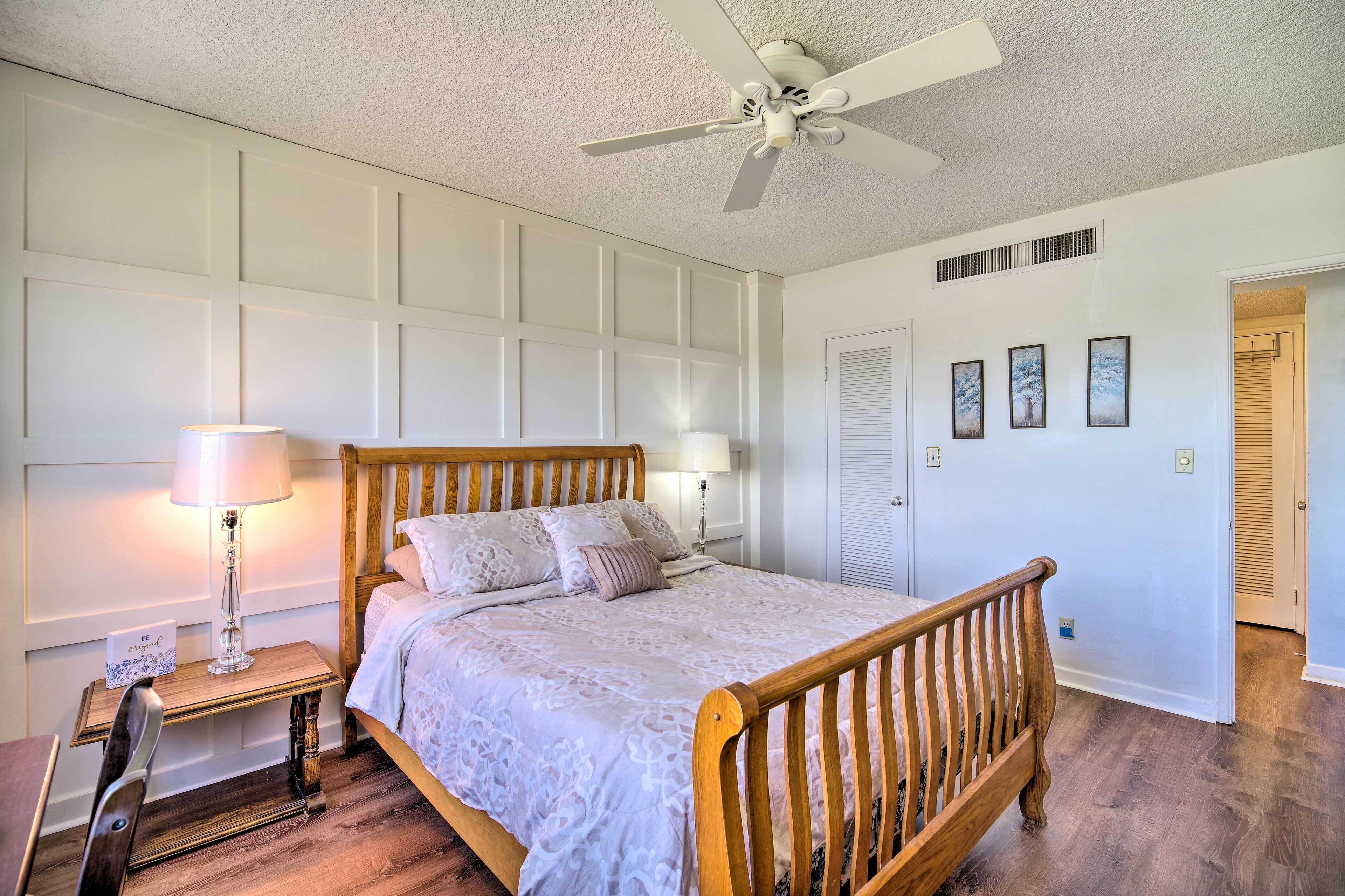 A bedroom with a wooden bed, two bedside lamps, a ceiling fan, and a door leading to another room.