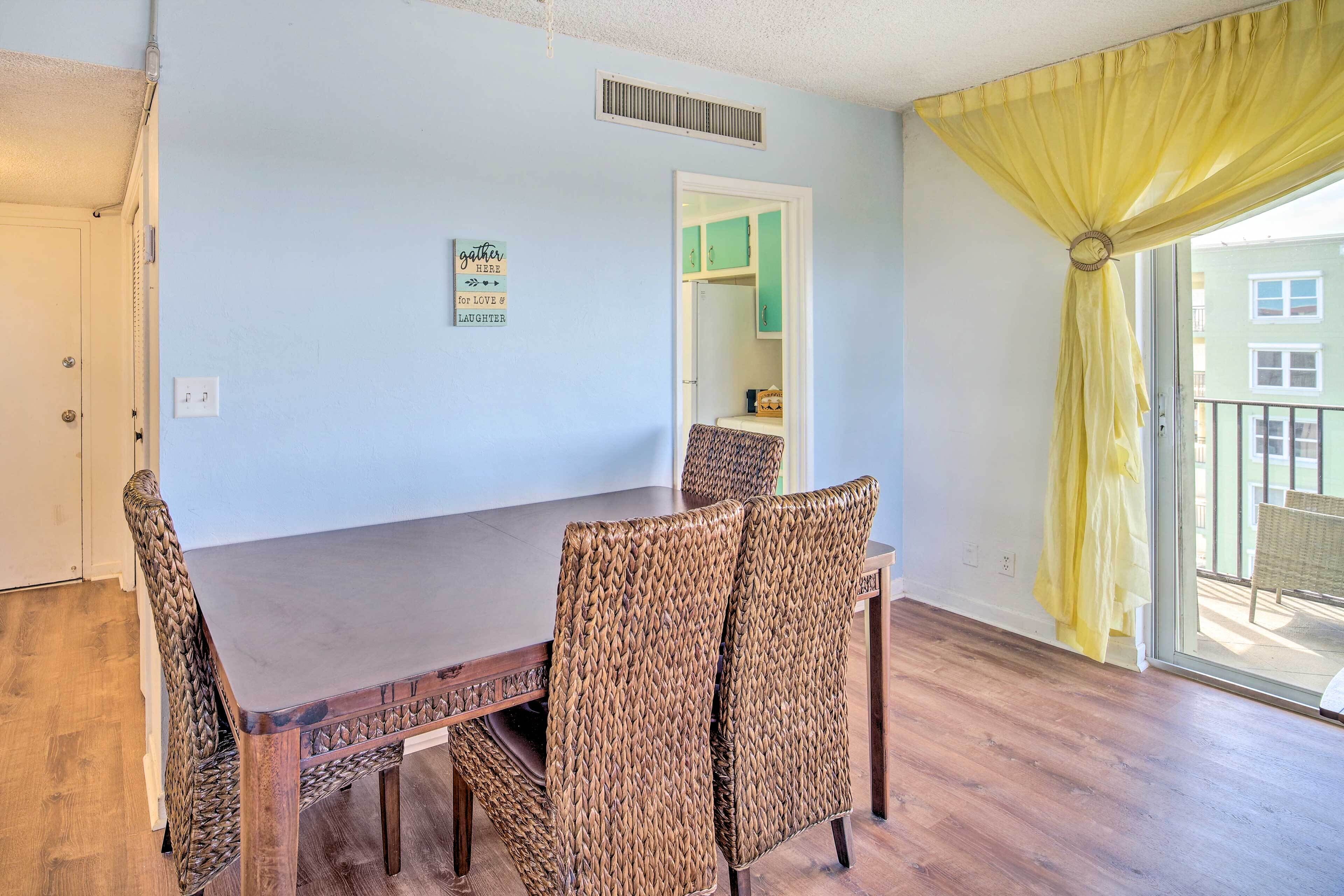 A dining area with a wooden table and wicker chairs, a light blue wall, a yellow curtain, and a view of a balcony.