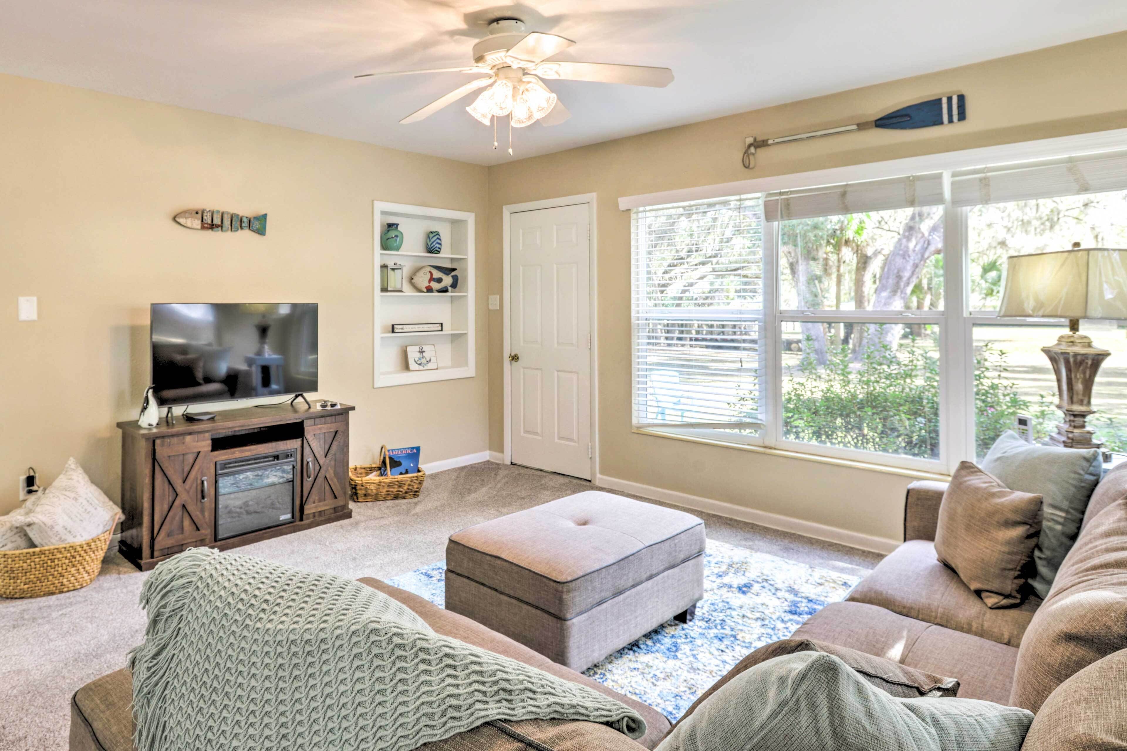 A living room with a television, ottoman, and sofa, a ceiling fan, and a window with blinds.