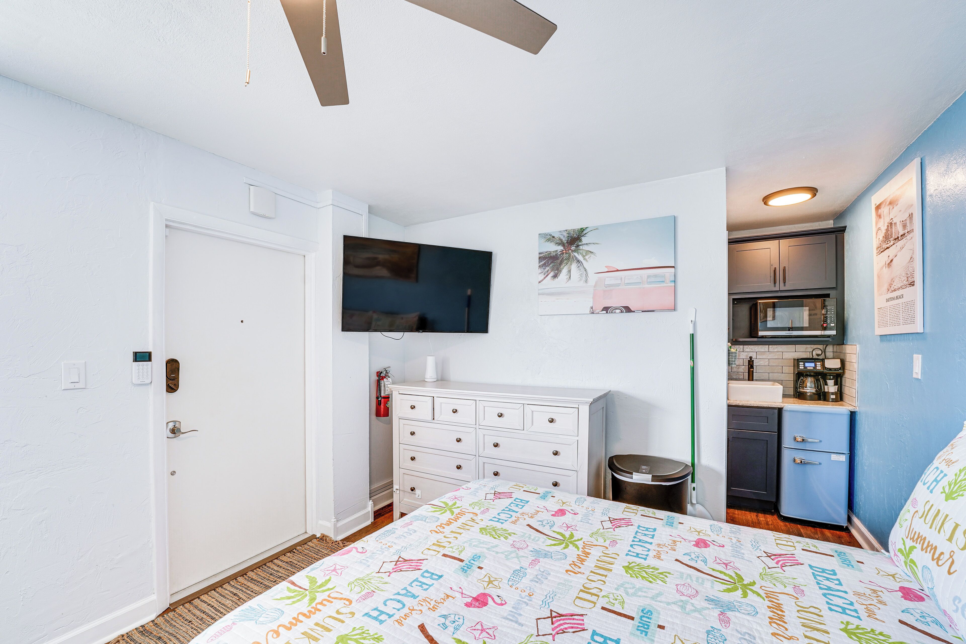 A compact bedroom with a bed, a dresser, a microwave, and a small kitchenette.