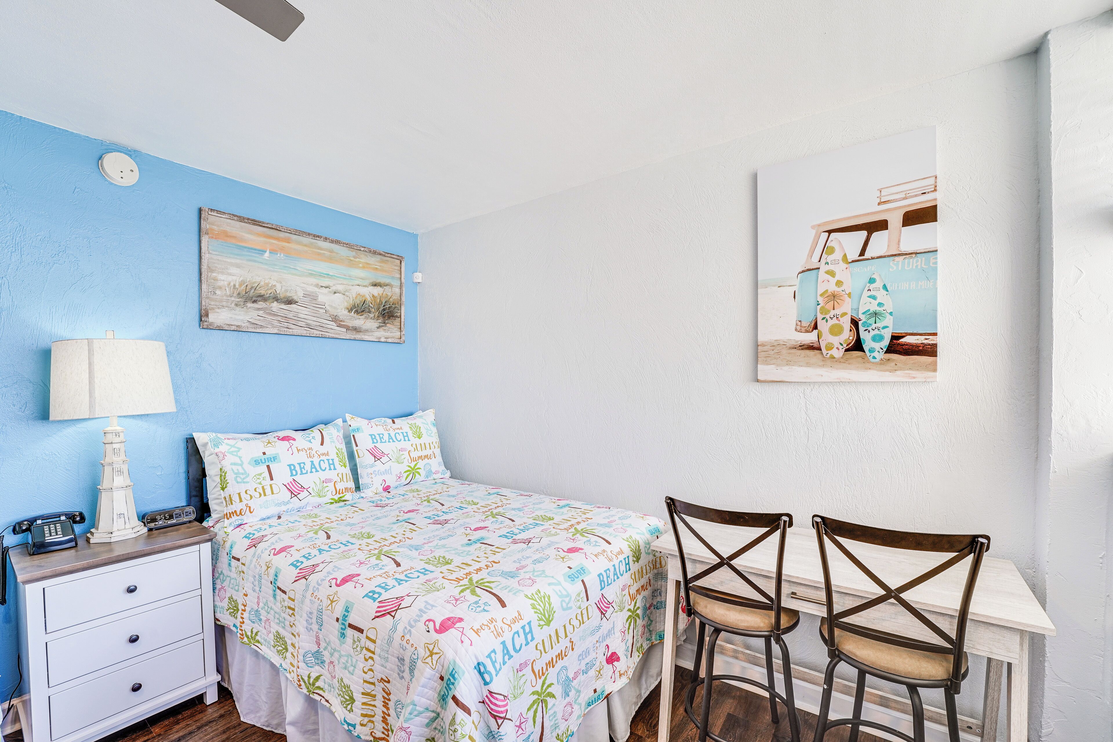 A bedroom with a bed, nightstand, lamp, and a painting of a beach scene.