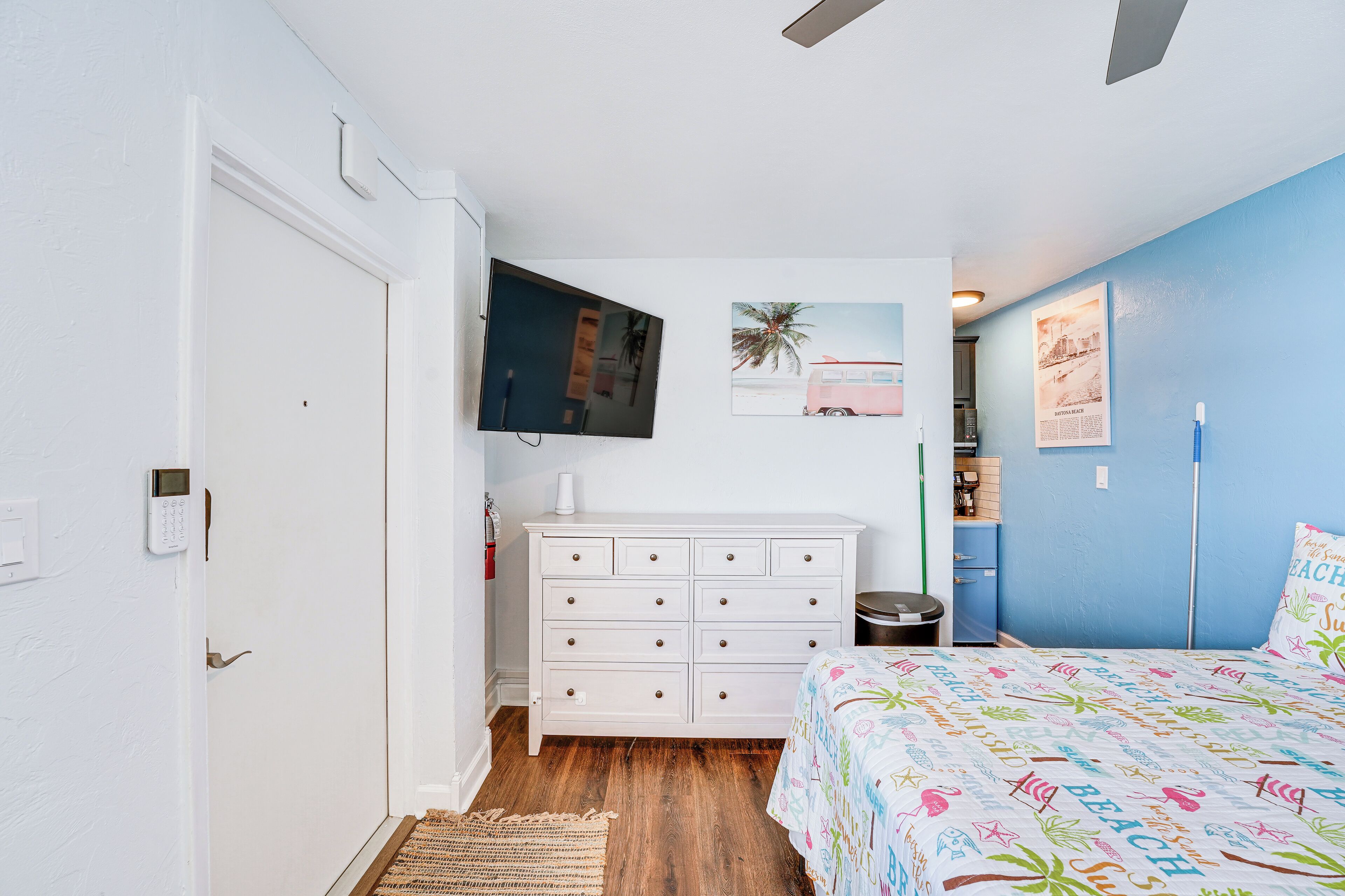 A bedroom with a white dresser, a bed with a colorful beach-themed bedspread, a mounted TV, and a wall with a palm tree and beach scene artwork.