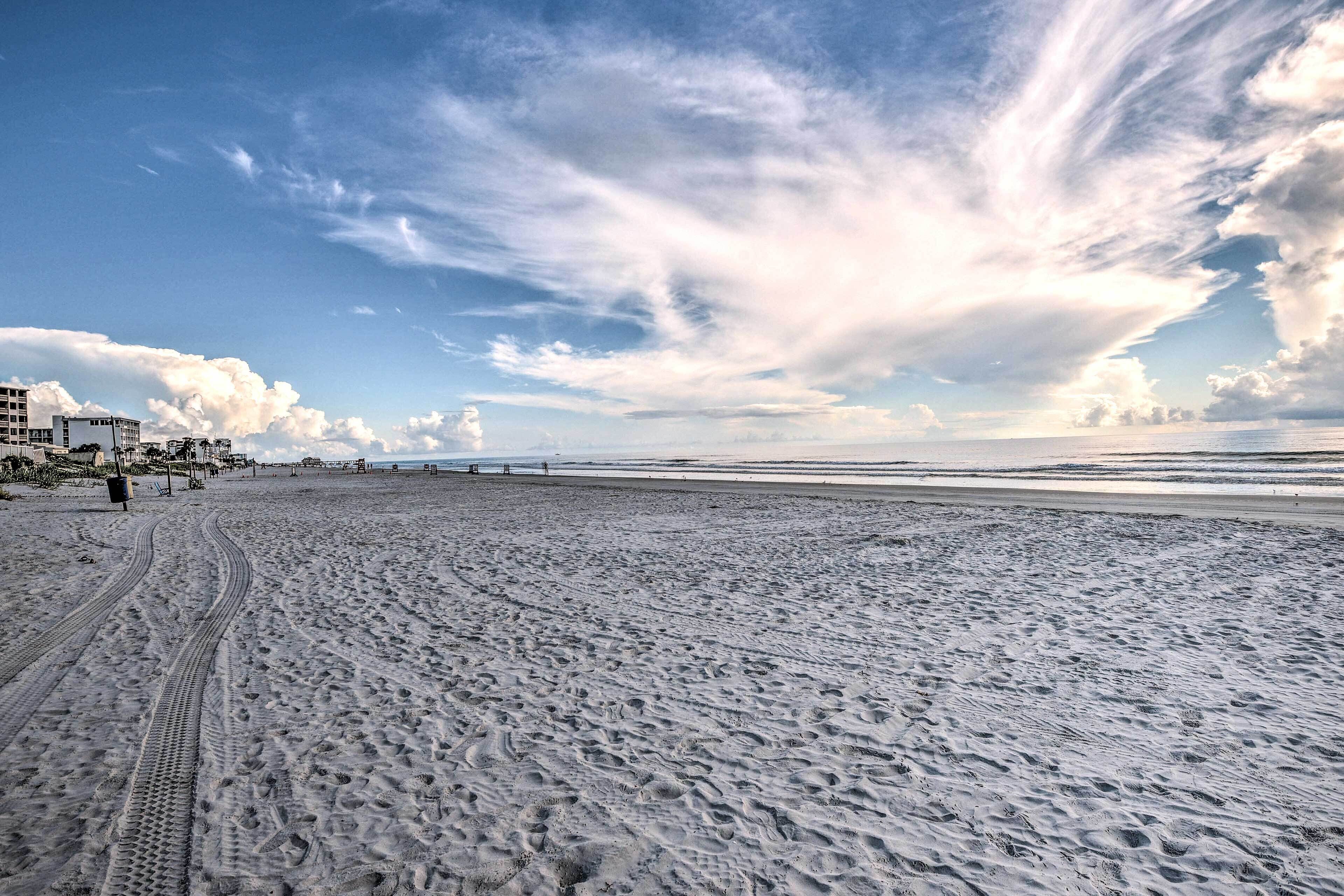 Une vaste plage de sable où l’on peut voir des traces de pneus, un ciel dégagé et des bâtiments qui se dessinent à l’horizon.