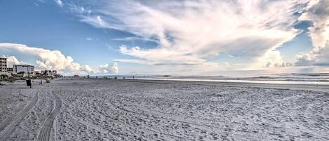 Une vaste plage de sable où l’on peut voir des traces de pneus, un ciel dégagé et des bâtiments qui se dessinent à l’horizon.