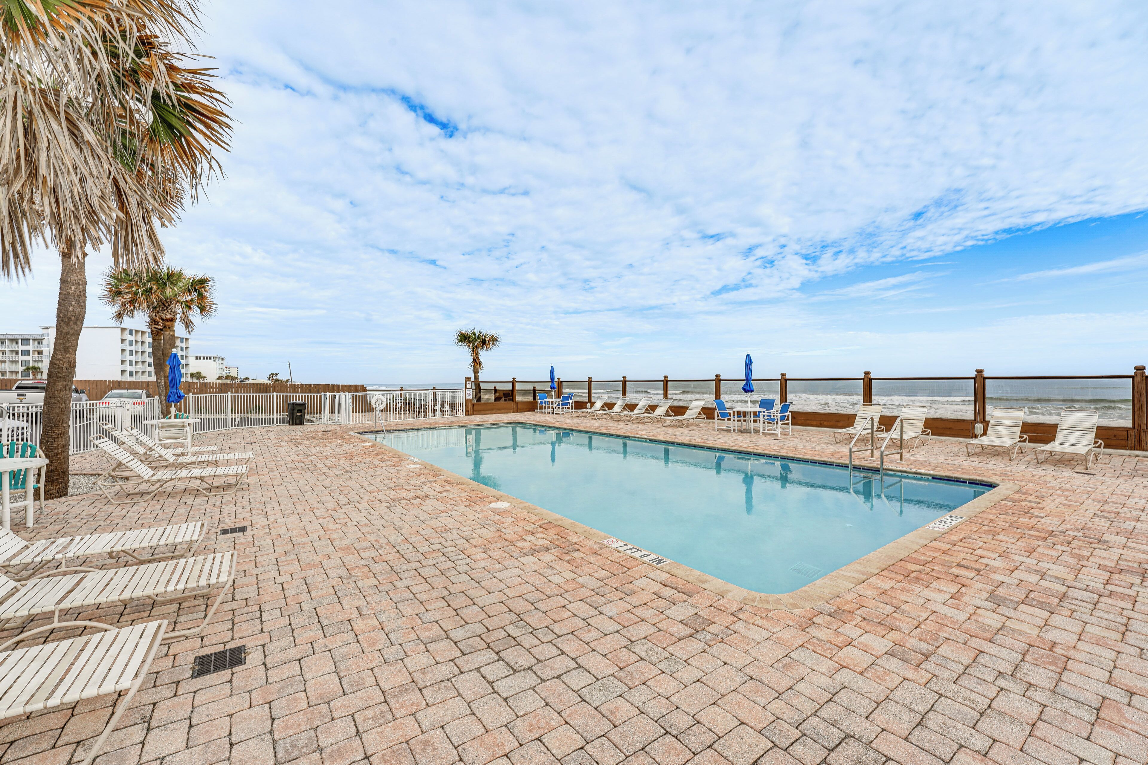 A pool area with lounge chairs, a wooden deck, and a view of the ocean.