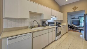 Une cuisine moderne avec des meubles blancs, un lave-vaisselle, une cuisinière et un évier. On aperçoit un téléviseur au fond.