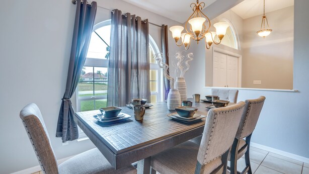 A dining area with a wooden table set for four, a chandelier, and a view of the outdoors through a window with curtains. - Private Yard & Pool: Stunning Cape Coral Getaway! (Cape Coral)