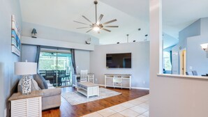 A modern living room with a ceiling fan, a TV mounted on the wall, a sofa, a coffee table, and a dining area with chairs and a table. - Private Yard & Pool: Stunning Cape Coral Getaway! (Cape Coral)