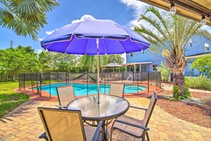 A pool area with a covered table and chairs, surrounded by a fence and palm trees.