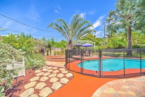 A swimming pool with a surrounding fence, a stone pathway, and lush greenery including palm trees.