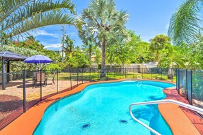 A swimming pool surrounded by a fence, with palm trees and a gazebo in the background.